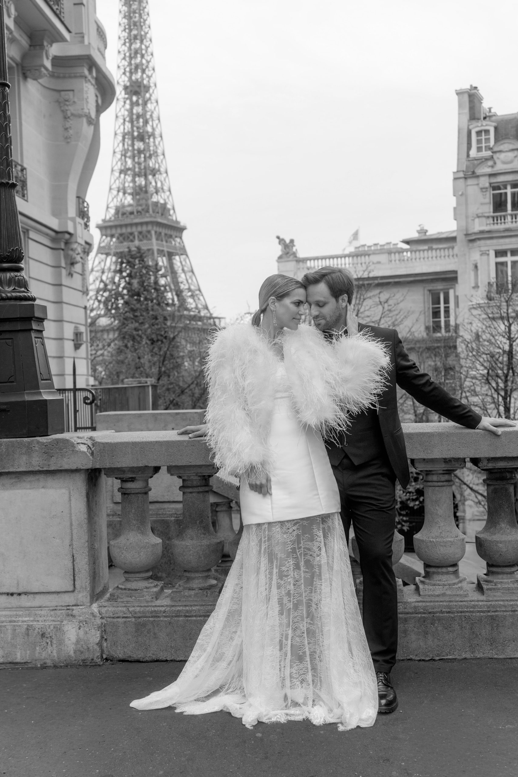 Couple de mariés élégants avenue Camœns à Paris avec vue sur la Tour Eiffel, mariée en veste à plumes blanches et robe moderne se regardant avec amour